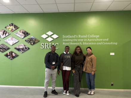 Four students posing with smiles in front of a green wall with the sign "Scotland's Rural College: Leading the way in Agriculture and Rural Research, Education and Consulting."